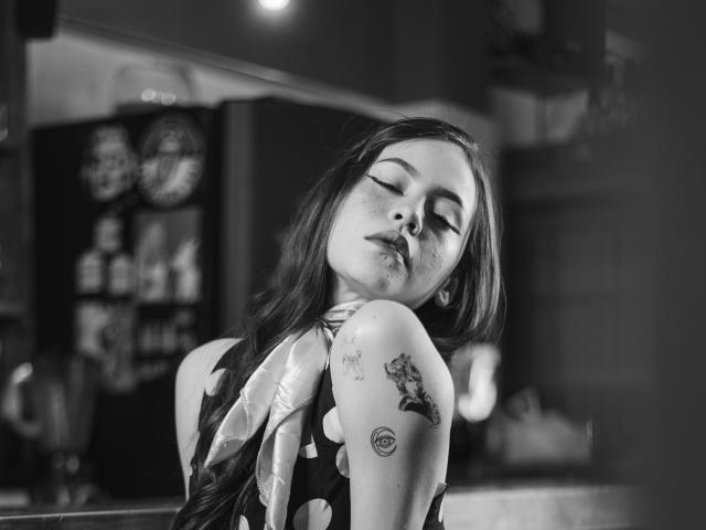 Jazmin Wells ha lunghi capelli scuri, gli occhi chiusi e dei tatuaggi sulla spalla e sul braccio. Indossa un vestito a pois e, in una foto in bianco e nero, ha un'espressione sensuale e leggermente accigliata..