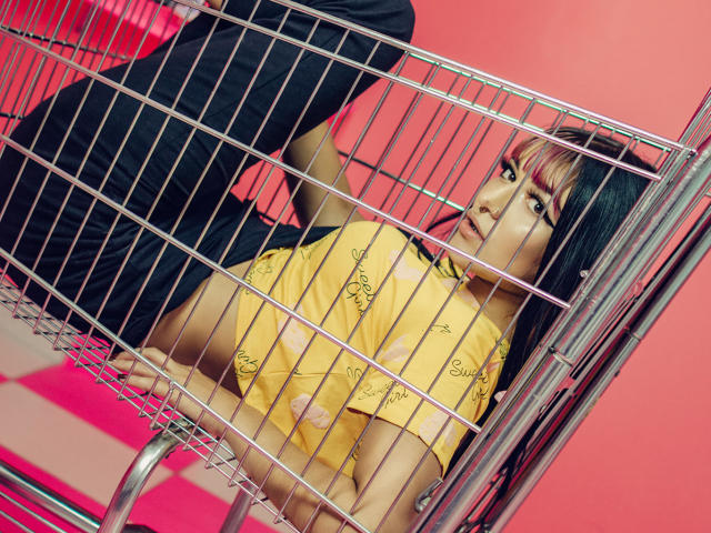 LindaWatsong's ass is in a metal cage, wearing a yellow shirt with a pattern, black pants, and pink highlights in her hair, looking up with a serious expression.