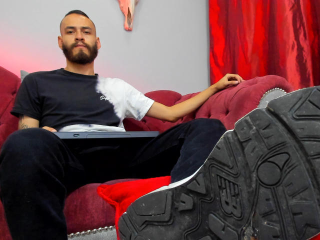 AndersX, com barba, vestindo uma camiseta preta e calças pretas, sentado em um sofá vermelho, com as pernas abertas, mostrando tênis Nike pretos, um laptop no colo, e um travesseiro vermelho atrás..