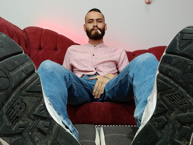 AndersX, con barba, una camicia rosa e jeans blu, seduto su un divano rosso con le gambe divaricate, mostra in primo piano degli stivali neri, consumati, e tiene in mano una sigaretta..