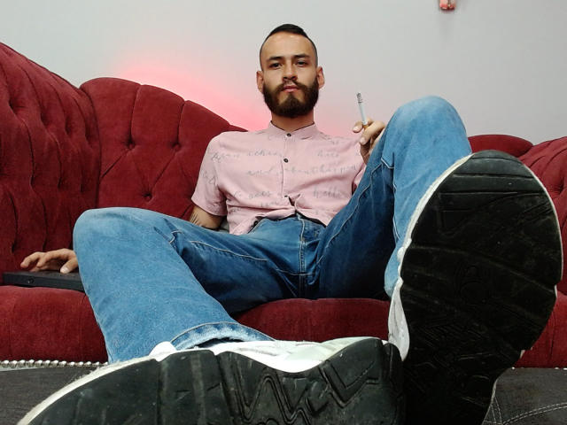 AndersX, avec une barbe, un chemisier rose et un jean bleu, tenant une cigarette, les jambes écartées, montrant des baskets blanches à semelles noires, assis sur un canapé en velours rouge..
