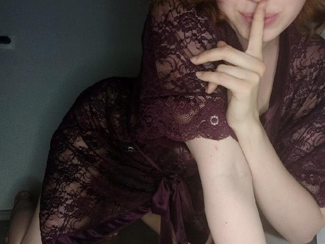 KiraNightly, vêtue d'une robe en dentelle violet foncé, se couvre la bouche avec son doigt, a une peau pâle et des cheveux roux, et ses seins sont légèrement visibles à travers la dentelle..