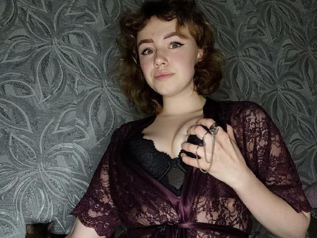 KiraNightly, con cabello castaño rizado, vestida con una bata púrpura de encaje transparente que deja ver un sujetador de encaje negro, sosteniendo un anillo y una cadena, sonriendo ligeramente y mirando a la cámara..