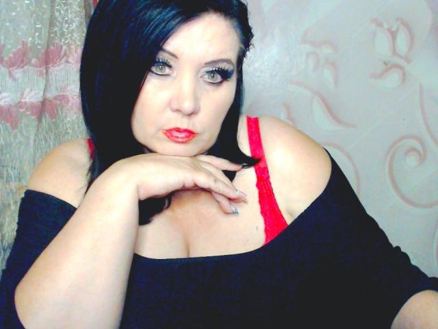 KamilaDream mit schwarzen Haaren, rotem Lippenstift und roten BH-Trägern, trägt ein schwarzes Top mit offenem Schulterbereich und legt ihr Kinn in die Hand, wobei ihre Brüste sichtbar sind..