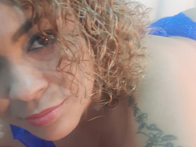 Solmary: Cabelos loiros cacheados, batom vermelho e tatuagem no ombro, vestindo renda azul, olhando para a câmera, mostrando um pouco dos seios. Luz suave, sorriso discreto..