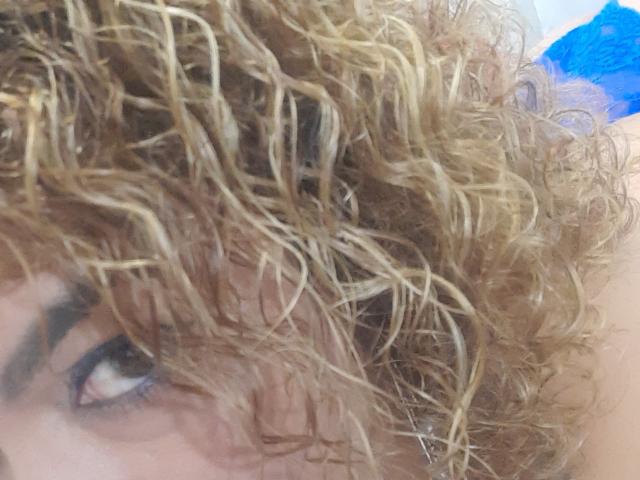 SolmaryHot hat lockiges, feuchtes, braunes Haar mit blonden Strähnen, wobei eine einzelne, dunkle Augenbraue und ein teilweise sichtbares Auge zu sehen sind. Im verschwommenen Hintergrund befindet sich ein blauer Stoff..