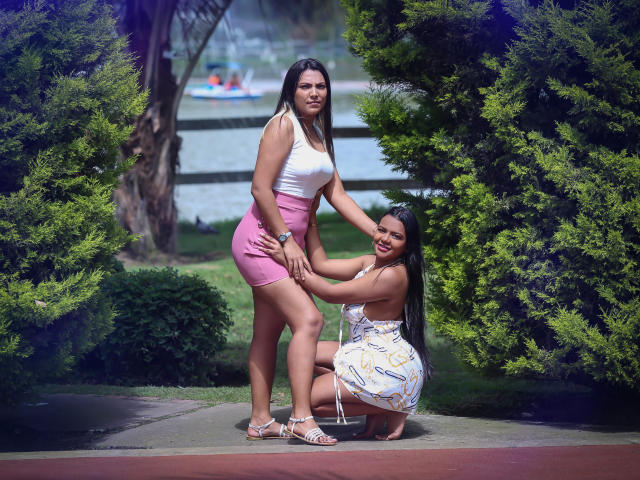 JadeAndDhanna, con cabello negro largo, vestida con una camiseta blanca y pantalones cortos rosas, está de pie, mientras que Dhanna está arrodillada, sosteniendo el trasero de JadeAndDhanna, y lleva un vestido blanco con estampado, en un parque soleado..