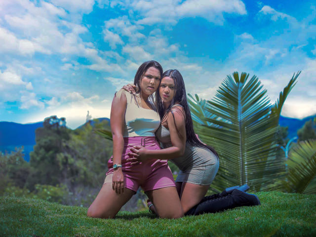 JadeAndDhanna met lang donker haar, knielend op het gras, Jade in roze shorts en een witte top, Dhanna in een grijze jurk, die Jade's billen vasthoudt, blauwe lucht, pluizige wolken, groot groen blad..