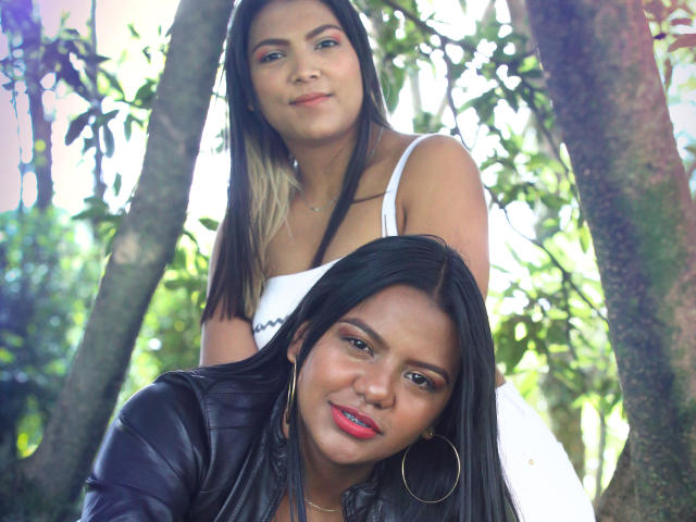 JadeAndDhanna, com longos cabelos pretos e batom vermelho, vestindo uma jaqueta de couro, em frente a Dhanna, com mechas loiras, vestindo uma blusa branca, em pé atrás dela..