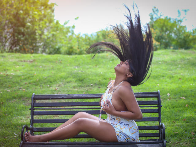 JadeAndDhanna com enormes seios negros à mostra, saindo de seu vestido branco, sentada em um banco preto, com um fundo de grama, luz do sol, olhos fechados, segurando seu grande traseiro, cabelos longos e pretos..