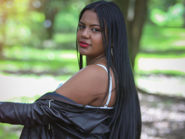 JadeAndDhanna con lunghi capelli neri, rossetto rosso, reggiseno bianco e giacca di pelle nera, che guarda oltre la spalla, mostrando la spalla scoperta e un leggero scollamento. Filigrana Boqboq..