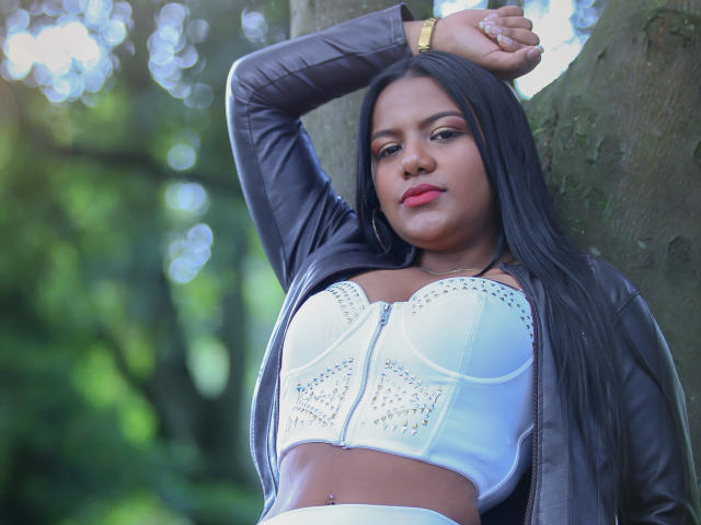 JadeAndDhanna, avec ses longs cheveux noirs, portant un soutien-gorge blanc orné de clous, une veste en cuir noir et une montre dorée, appuyée contre un arbre, les bras levés, montrant ses seins..