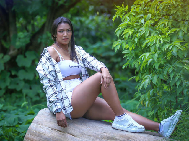 JadeAndDhanna sitting on a rock, wearing a white sports bra, plaid shirt, high-waisted shorts, and white sneakers, with her legs spread, showing her ass. Green leafy background.