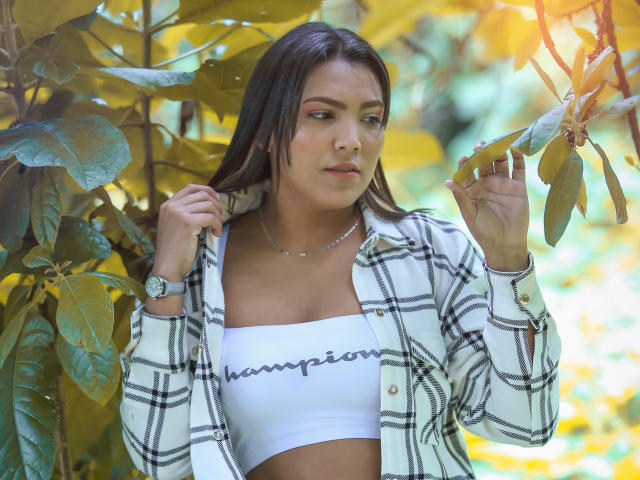 JadeAndDhanna mit mittelgroßer Brust, trägt ein weißes Crop-Top, ein kariertes Hemd und eine Uhr, hält ein Blatt, Sonnenlicht fällt durch, grüne und gelbe Blätter im Hintergrund, ernster Gesichtsausdruck, mittelbraunes, halblanges Haar..