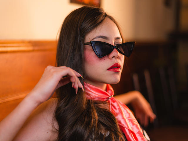 Jazmin Wells, con lunghi capelli castani, occhiali da sole neri a forma di occhi di gatto, rossetto rosso e una sciarpa rosa, che si tocca i capelli, guardando di lato, in un ambiente caldo e con una luce soffusa..