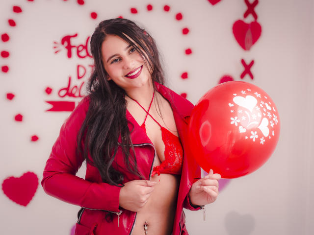 LeilaEvanns, con lunghi capelli neri, labbra rosse e un sorriso, indossa un reggiseno e una giacca di pizzo rossi, tiene in mano un palloncino rosso con cuori bianchi, mostrando il seno..
