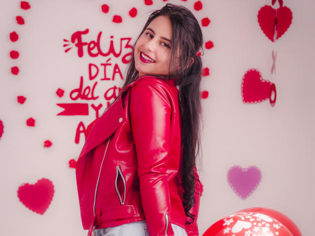 LeilaEvanns mit langen, dunklen Haaren, einer roten Lederjacke und blauen Jeans, lächelt in die Kamera und zeigt ihren roten Lippenstift und ihren attraktiven Hintern. Im Hintergrund sind rote Herzen und rosa Pompons zu sehen..