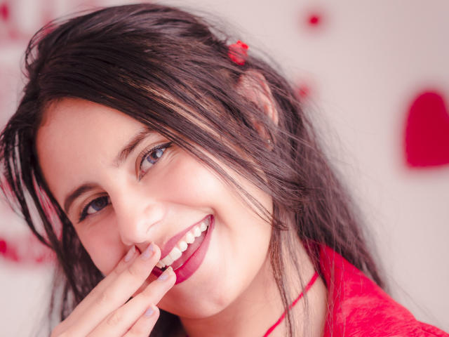 LeilaEvanns, com cabelo castanho escuro, olhos azuis e batom vermelho, sorrindo, tocando seus lábios vermelhos com o dedo, vestindo uma blusa vermelha de renda, parecendo sexy e adorável..