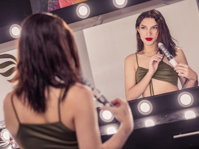 LaurynsKalii, com cabelo escuro, blusa verde cropped, segurando um microfone, olhando para si mesma em um espelho com luzes redondas, batom vermelho e um leve sorriso. Seios visíveis..