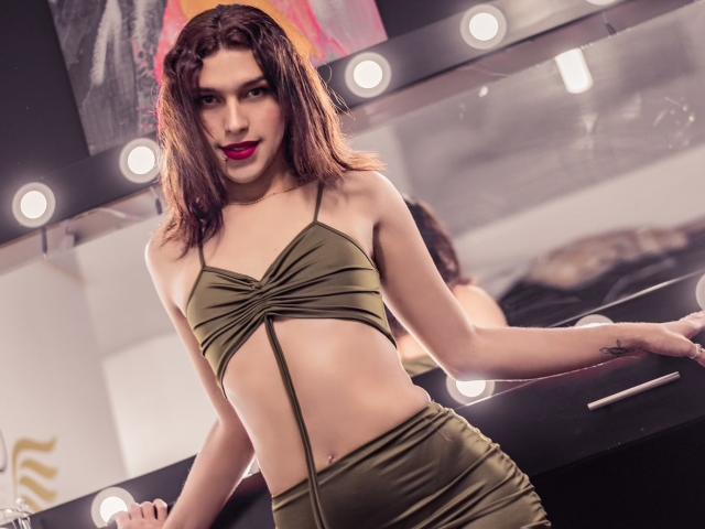 LaurynsKalii, avec ses cheveux bruns ondulés, porte un haut vert olive, découpé et sans bretelles, ainsi qu'une jupe assortie à taille haute. Elle est appuyée contre un comptoir noir, avec du rouge à lèvres, un miroir et des lumières..
