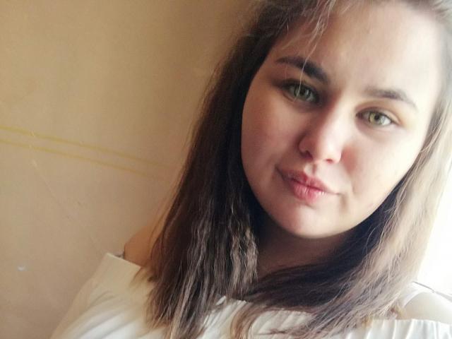 KaidoKun tiene ojos verdes, cabello castaño de largo medio, y una ligera expresión de puchero con labios rosados, vistiendo una blusa blanca, en un selfie con una iluminación suave y ligeramente borroso..