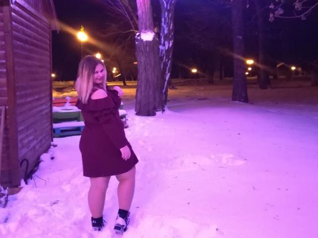 KaidoKun steht im Schnee, trägt ein rotes Kleid mit Ausschnitten an den Schultern, schwarze Schuhe und langes blondes Haar, unter violetten Nachtlichtern, neben einem Holzschuppen..