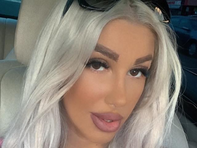 TanyaMonroe has platinum blonde hair, thick eyebrows, long lashes, and pink lipstick. Her brown eyes are slightly open, and she's wearing black sunglasses on her head.