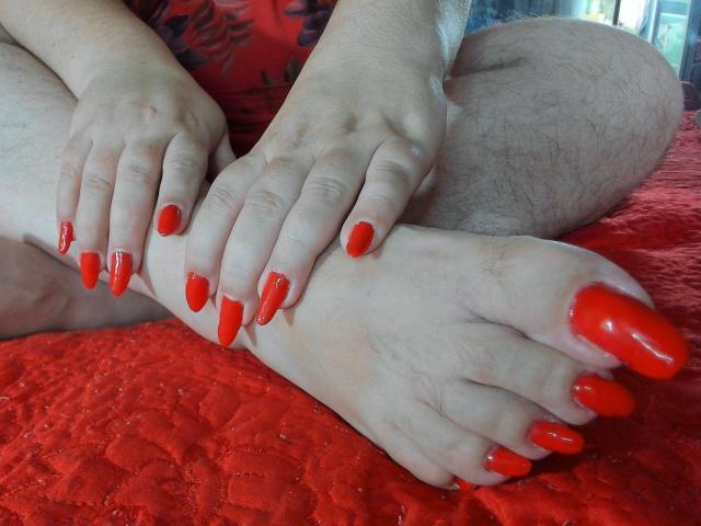 KaryQueen's feet with bright red, long, sharp nails, resting on a red quilted bed. Her hands with matching nails are gently touching her feet. Her hairy legs are visible.