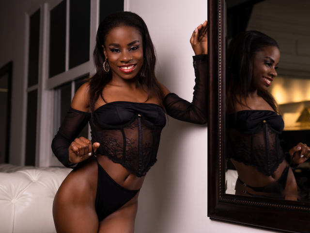 MiaHopson with dark skin, wearing a black lace top and black panties, standing, smiling, hand on mirror, reflection showing her tits and ass, long brown hair, hoop earrings.