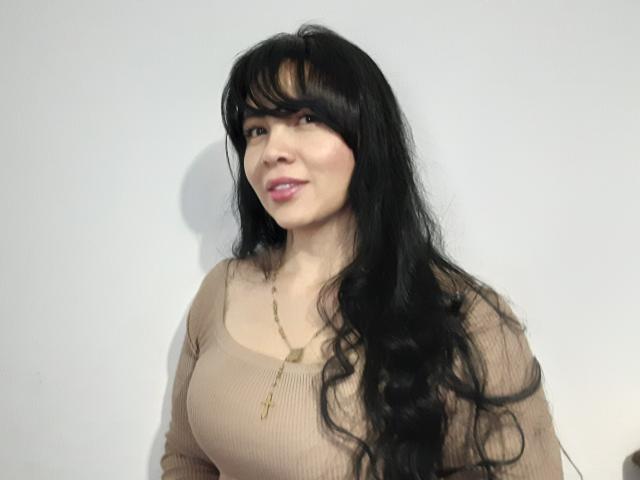 ArianaCoollins, com longos cabelos pretos ondulados, vestindo uma blusa bege texturizada, mostrando seus seios grandes, lábios rosados e um colar de ouro, sorrindo suavemente..
