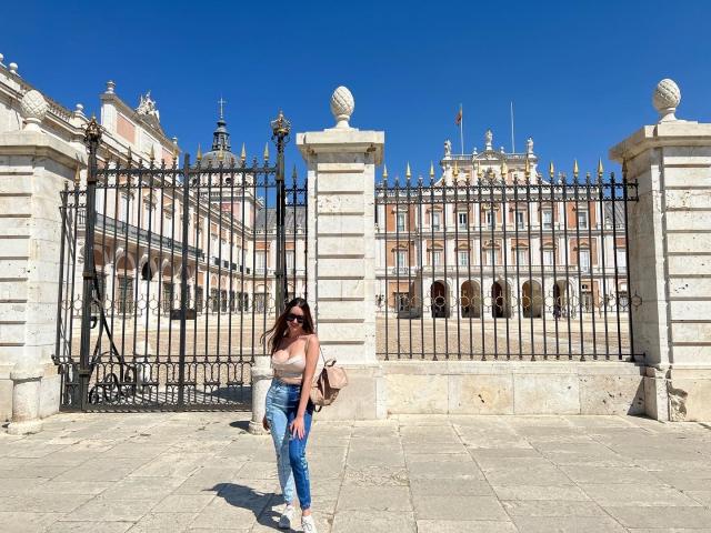 SusannaTaylor has big tits, wearing a pink top, blue jeans, and sunglasses, standing in front of a white, ornate, iron gate, with a clear blue sky.