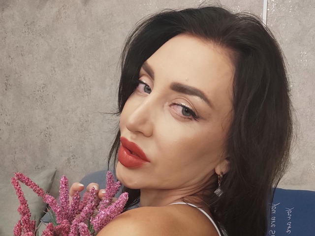 SimoniGrace hat dunkles, welliges Haar, roten Lippenstift und hält rosa Seeanemonen in der Nähe ihrer Brüste. Sie schaut über ihre Schulter mit einem leichten, verführerischen Lächeln..