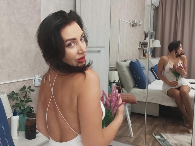 SimoniGrace, avec ses cheveux brun foncé, regardant par-dessus son épaule, portant une robe blanche sans dos, tenant des fleurs roses, son postérieur exposé, reflété dans un miroir, assise sur un canapé..