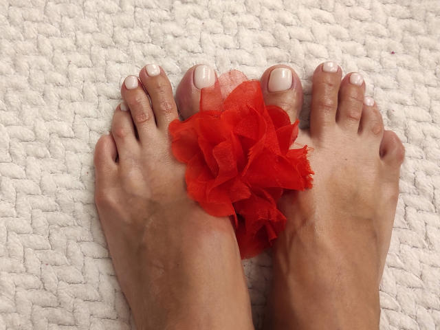 SimoniGraces Zehen mit weiß lackierten Nägeln, ein rotes Blumen-Haargummi an ihren Füßen, stehend auf einem strukturierten weißen Teppich..