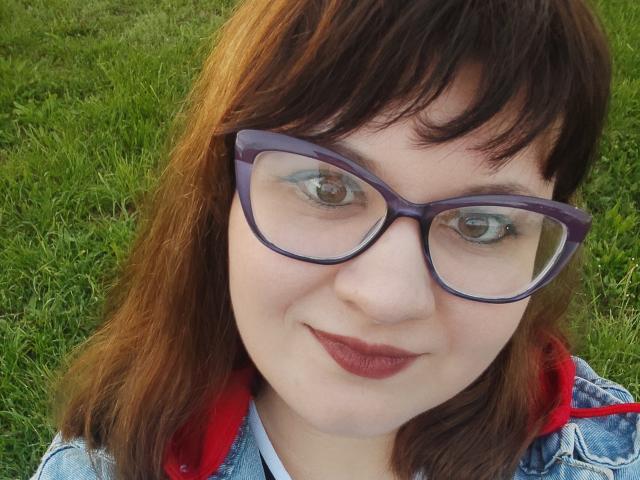 AlisaMi has brown hair, purple glasses, and red lipstick. She's wearing a denim jacket with a red hood. Her ass is not visible. Her tits are small.