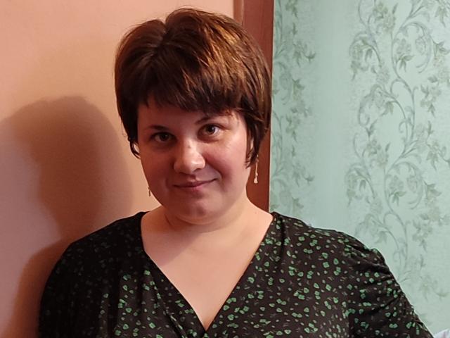AlisaMi heeft kort bruin haar, een lichte huid en kleine borsten. Ze draagt een zwarte jurk met groene patronen, glimlacht licht en staat tegen een roze muur..