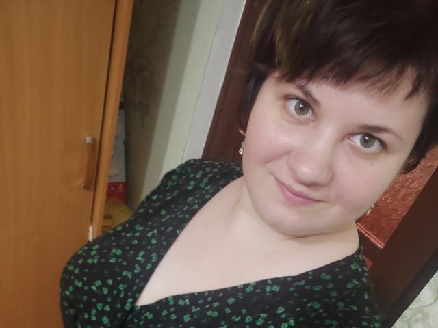 Alisa, com cabelo castanho curto, olhos verdes e um leve sorriso, vestindo um vestido preto com padrões verdes, mostrando seus seios. Sua pele é clara e ela está em uma porta..