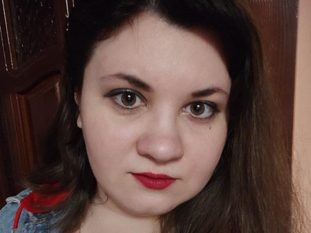 El rostro de AlisaMi, ojos marrones, lápiz labial rojo, ligera sonrisa, piel clara, cabello marrón oscuro, vestida con jeans y tirantes rojos, puerta de madera en el fondo..