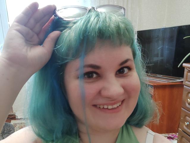 AlisaMi, com cabelo verde-azulado, sorrindo, segurando óculos na cabeça, com uma mecha azul no cabelo, manga branca, nariz rosa, pele clara, olhos castanhos, em um quarto aconchegante..