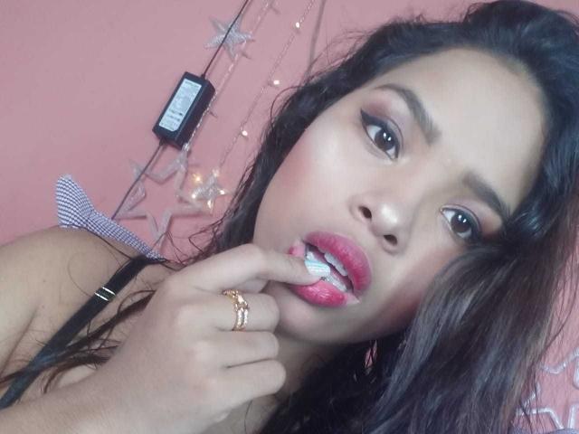 Liama69: Een meisje met donker haar, roze lippenstift en een ring, die op een tandenborstel zuigt en witte tandpasta op haar roze lippen laat zien, gekleed in een zwarte bandeau, in een roze kamer..