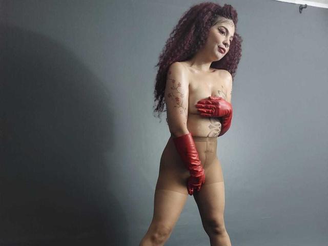 AndiChin with curly red hair, red gloves, covering her tits with her hands, wearing sheer pantyhose, standing against a dark grey wall, showing tattoos on her right arm.
