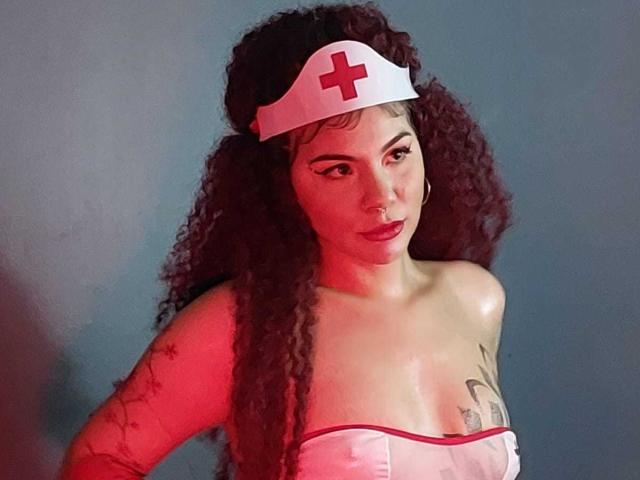 AndiChin, avec ses cheveux bruns bouclés, porte un bonnet d'infirmière blanc avec une croix rouge, un haut blanc sans bretelles, laissant entrevoir de petites seins et des tatouages, et regarde l'appareil photo sous un éclairage rouge..
