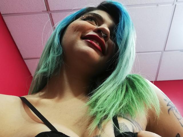 AndiChin, con capelli blu-verdi, piercing al naso, rossetto rosso, sorridente, indossa un reggiseno nero, ha tatuaggi sul braccio, guarda in basso, sullo sfondo si vedono il soffitto e una parete rosa..