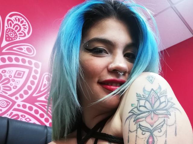 AndiChin met blauw haar, zwarte eyeliner, rode lipstick, een neusring en een tatoeage op de rechterarm met een roze lotusmotief. AndiChin's borsten zijn gedeeltelijk zichtbaar..