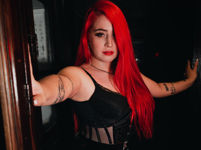 AprilDoel with bright red hair, black lace-up corset, and tattoos on arms, extending arms to frame doorway, red lipstick, and confident expression, in dark, moody setting.
