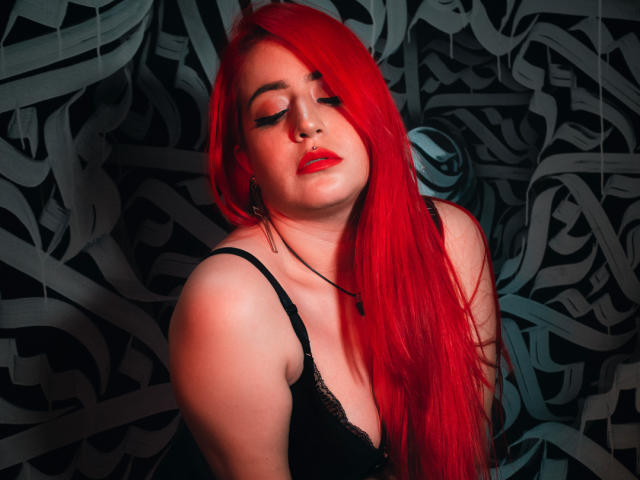 April Doel, con el cabello rojo brillante, ojos cerrados, vistiendo un sujetador de encaje negro, lápiz labial rojo, un collar y aretes, tiene una apariencia sexy y segura, mostrando sus senos y un rostro lindo y atractivo..