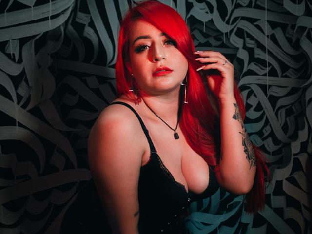 April Doel, avec ses cheveux roux éclatants, portant de la lingerie en dentelle noire, montrant ses seins, son bras tatoué et son rouge à lèvres rouge, sur un fond sombre et à motifs, regardant de manière séduisante l'appareil photo..
