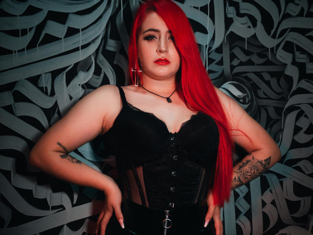 April Doel, con cabello rojo brillante, un corpiño de encaje negro y tatuajes, con las manos en las caderas, un collar con una cruz negra, lápiz labial rojo, de pie con confianza, mostrando sus pechos y tatuajes..