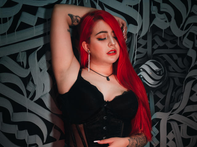 April Doel, con cabello rojo brillante, un corpiño de encaje negro con cordones y los senos levantados, con un brazo levantado, ojos cerrados, lápiz labial rojo, tatuajes y aretes, frente a una pared oscura con un patrón abstracto..
