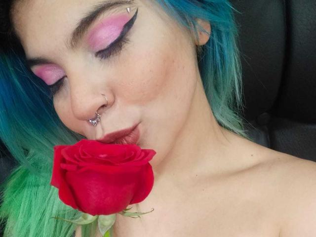 AndiChin, avec ses cheveux bleu-vert, son fard à paupières rose, son anneau au nez et son piercing aux lèvres, embrasse délicatement une rose rouge en la pressant contre ses lèvres, ses épaules dénudées, un siège de voiture noir en arrière-plan..
